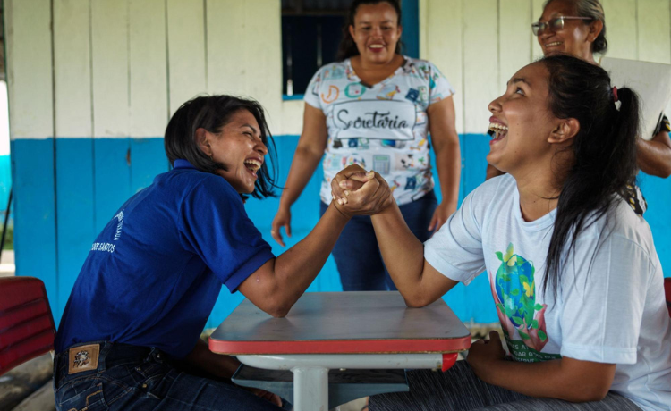 Two women sit at a table and arm wrestle, laughing; two more women watch on smiling in the background